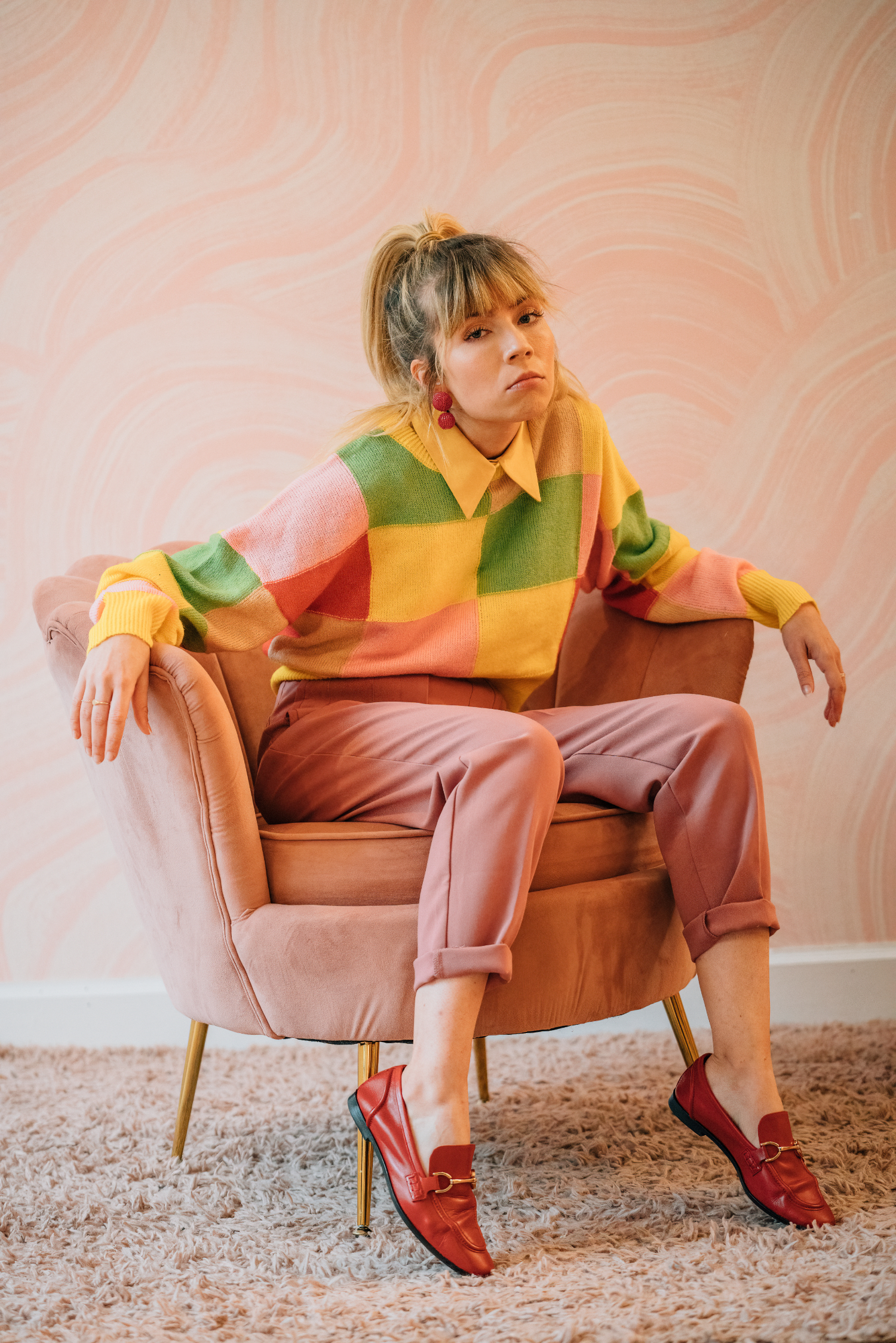Woman in colorful sweater and pink pants sitting in a pink chair against a pastel patterned wall.