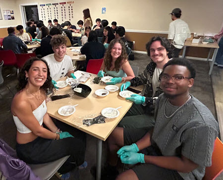 A crowd of students sit around tables with bowls, rice, plastic wrap, and gloved hands ready to make rice balls