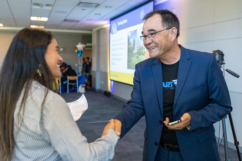 A woman in a striped blouse with long, dark hair shakes hands with a smiling man with short, salt-and-pepper hair. He wears a black t-shirt with the SpartUp logo under a blue suit.