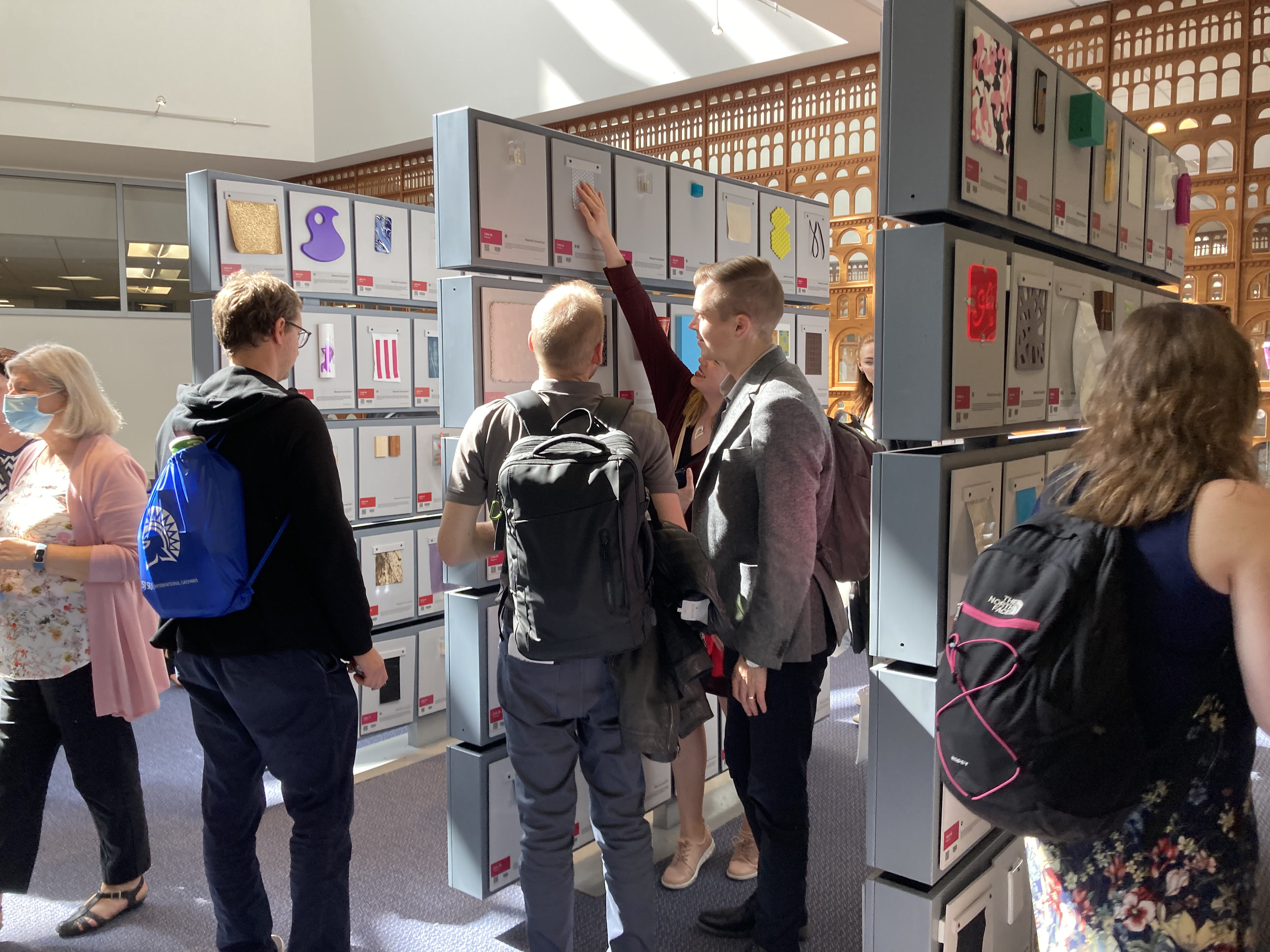 Three male students and one female stand in front of a panel of squares, each with a 4' scrap of plastic, fabric, or other material in SJSU's Materials Library. A staff member with gray hair and a pink sweater walks away in the background, while a woman with a background walks to the right in the foreground.