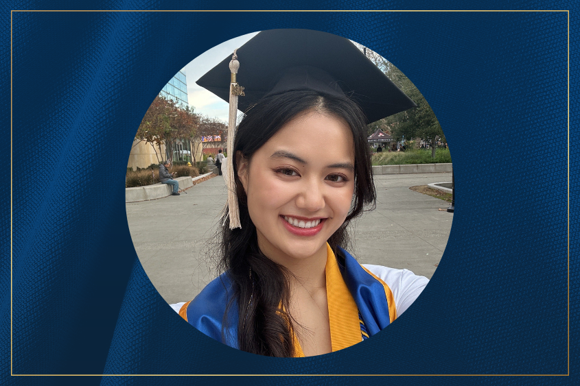 A graduate with long hair to the side wearing a black cap and blue and gold gown poses outdoors, celebrating their academic achievement amidst a festive backdrop.