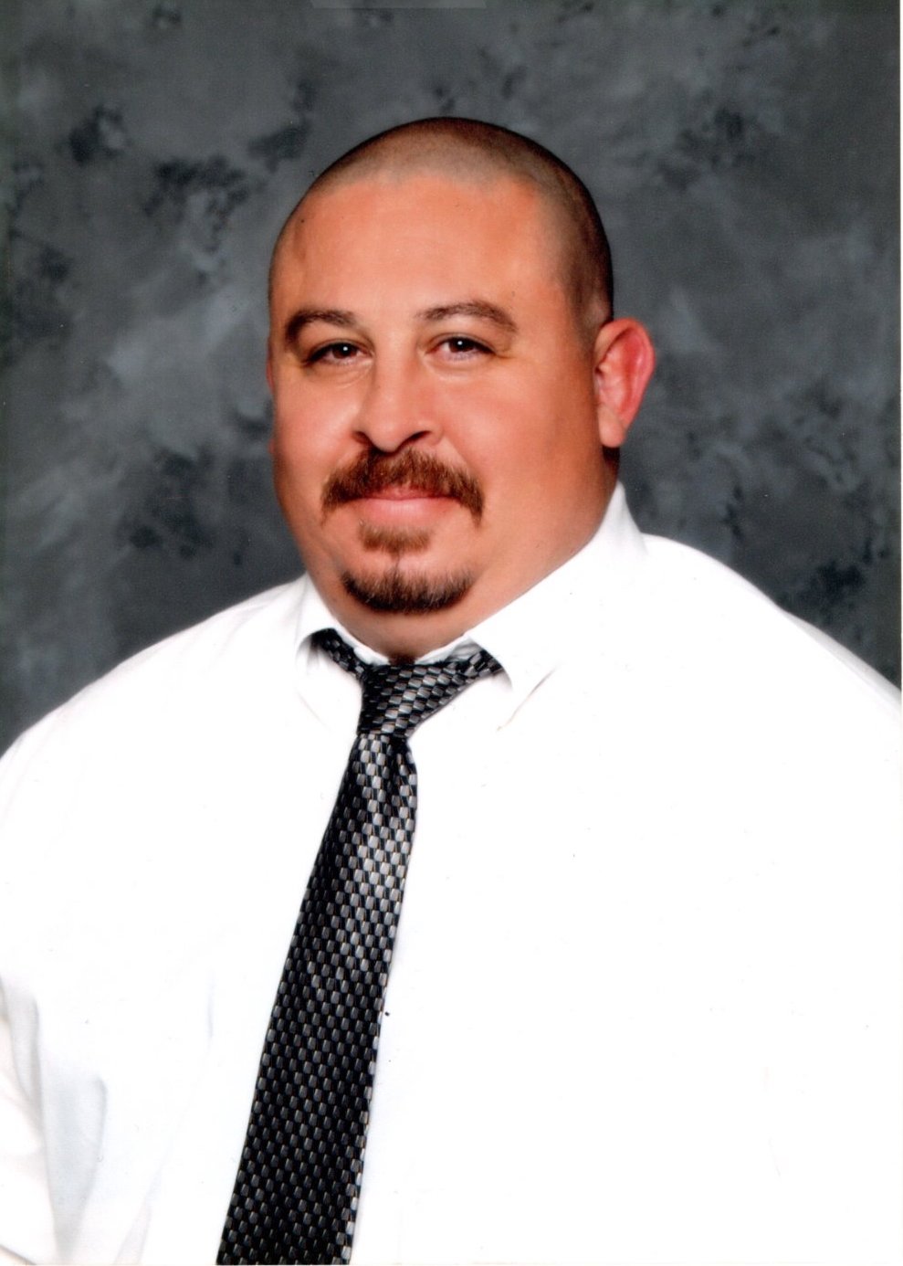 Armando Hispanic male with buzzed, brown hair and a brown moustache, wearing a white button-down and black tie, smiling in front of a black background.