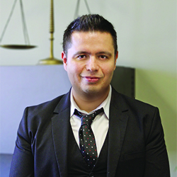 Claudio Hispanic male with short, black hair, wearing a black suit with a white, button-down shirt and a white spotted, black tie, Smiling in front of brass-colored justice scales.