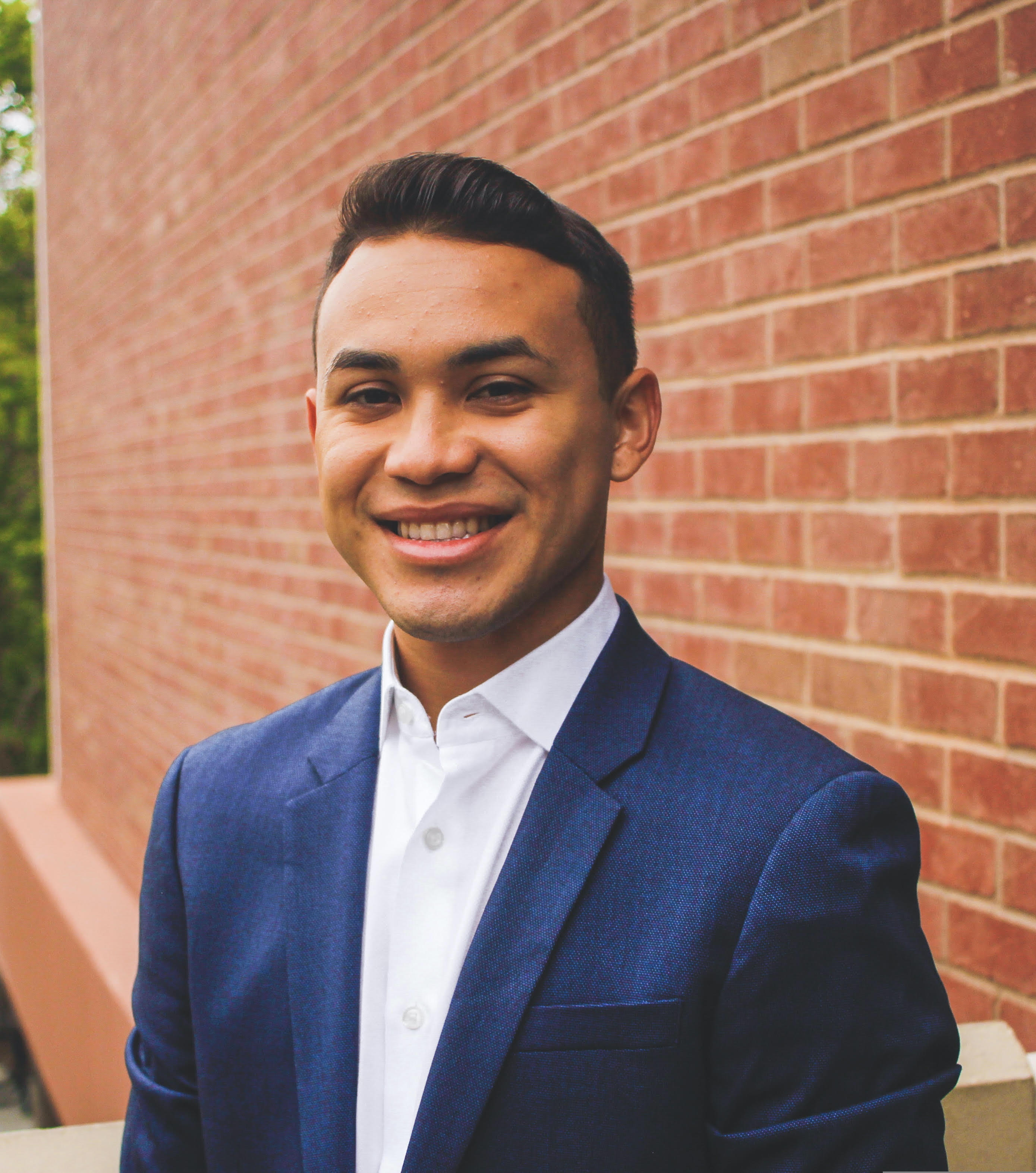 DC Hispanic male with short, black hair, wearing a blue suit jacket and white button down, smiling in front of a red brick wall.