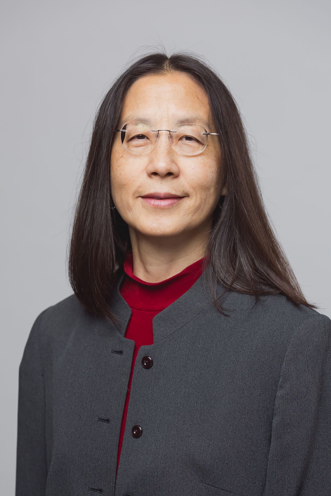 marina-hsieh Asian woman with long, black hair, with rounded silver glasses, wearing a gray suit jacket and red shirt, smiling in front of a gray background.