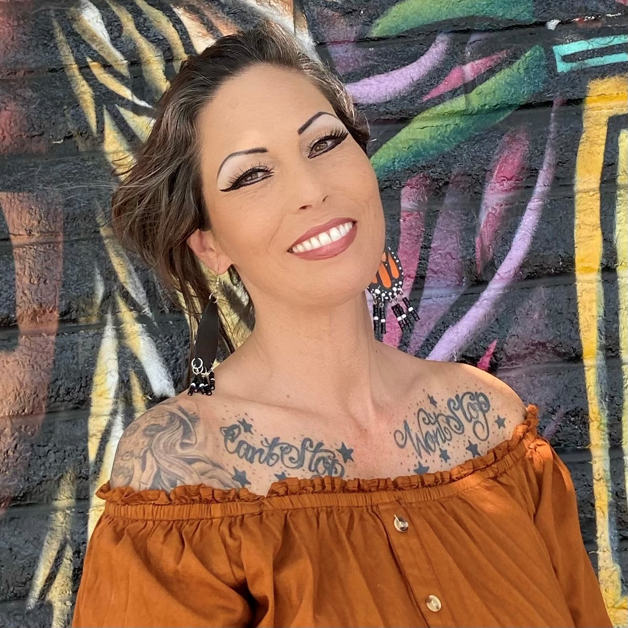 WP Hispanic woman with tattoos near her shouler, with medium length brown hair, wearing an orange shirt, smiling in front of grafitied wall.