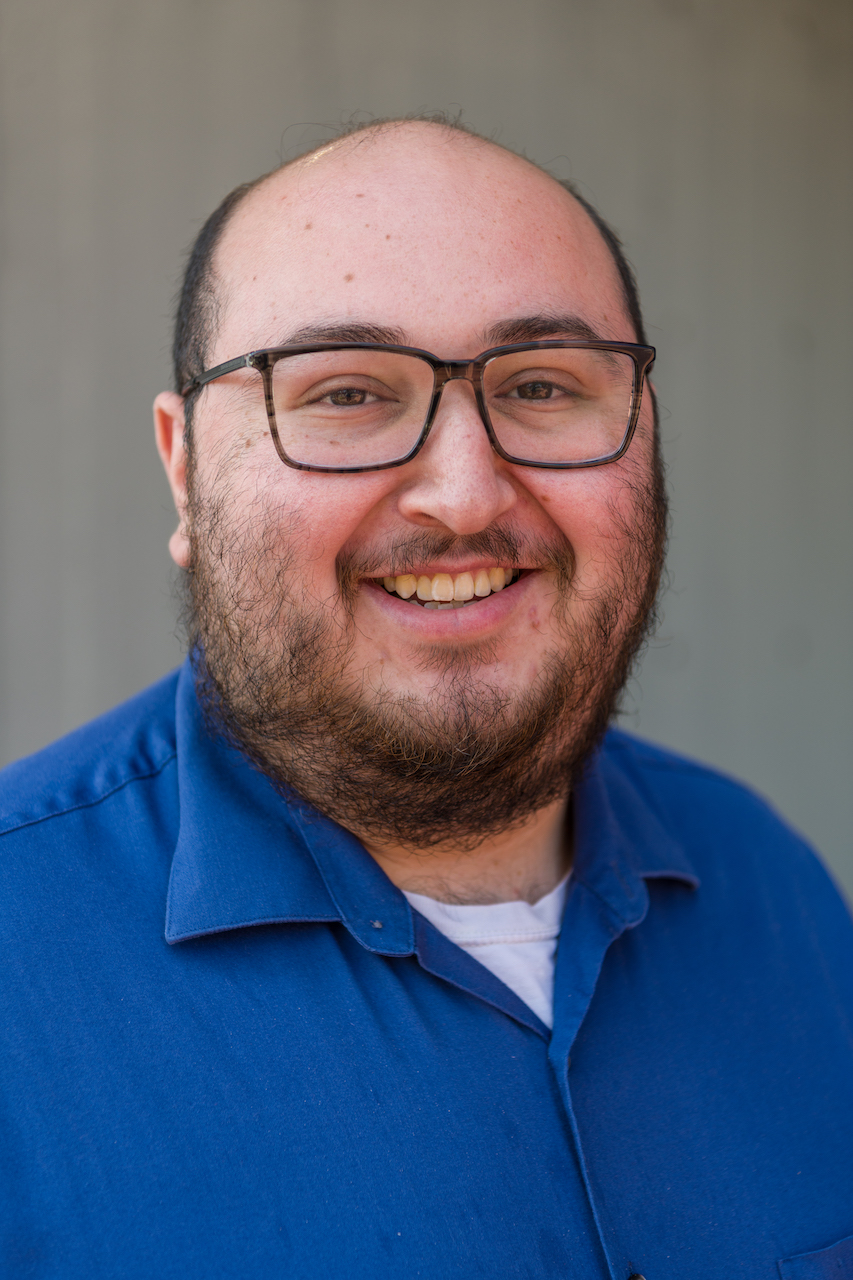 jesse-mejia Male in a blue, collared shirt, wearing glasses, smiling in front of a gray background.
